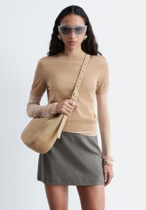 Femme aux longs cheveux noirs portant un haut beige à manches courtes sur des manches longues transparentes, une jupe grise, de grandes lunettes de soleil transparentes, un sac à bandoulière beige et des boucles d'oreilles argentées.