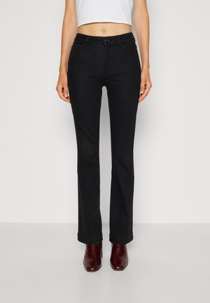 Kobieta ubrana w czarne jeansy bootcut, biały krótki top i ciemnoczerwone botki, stoi na tle prostego jasnego tła.