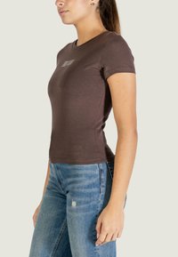 Hnědé přiléhavé tričko s krátkými rukávy, kulatým výstřihem a malým potiskem na hrudi. Kombinováno se světle modrými roztrhanými džínami.