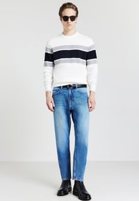 Maglione a righe bianche e blu navy, jeans blu con sfumature sbiadite e stivali di pelle neri. Il modello indossa occhiali da sole scuri e si trova in piedi su uno sfondo semplice.
