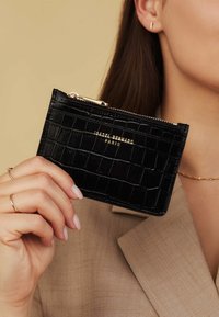 Porte-cartes en simili-crocodile noir avec une fermeture éclair dorée et un logo. Présente une surface texturée et une forme rectangulaire.