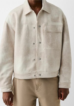 Homme portant une veste en daim beige clair avec des boutons pression et une poche poitrine à rabat, associée à un pantalon kaki sur un fond uni.