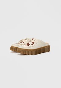 Scarpe slip-on con tomaia in pelle beige, dettaglio a macchie di mucca marroni e bianche, e suola in gomma marrone con spessore rigido, presentate su uno sfondo bianco.