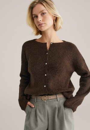 Blonde woman wearing gold hoop earrings, brown fuzzy button-up sweater, gray high-waisted pleated pants, and brown belt with gold buckle.