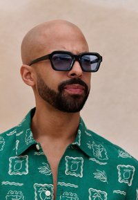 Black, angular sunglasses with dark lenses. Worn with a green, patterned shirt featuring white leaf and line designs. Smooth, bald head.