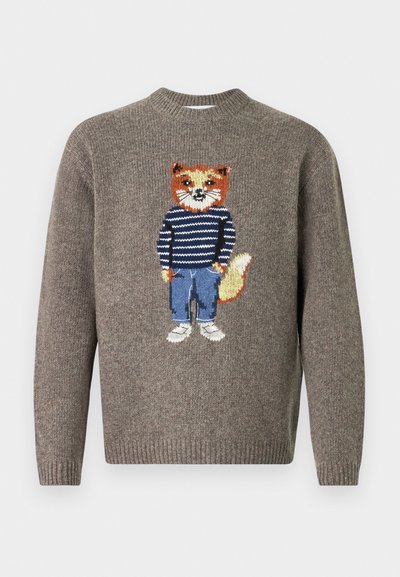 Brūns adījums džemperis ar izšūtu lapsu, kas valkā svītrainu džemperi, ziliem džinsiem un baltām kurpēm, priekšpusē.