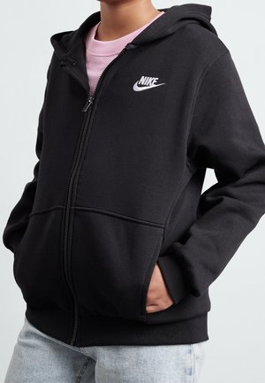 Osoba ubrana w czarną bluzę Nike z zamkiem błyskawicznym, kapturem i przednimi kieszeniami, połączoną z jasnoniebieskimi dżinsami i różową koszulką pod spodem.