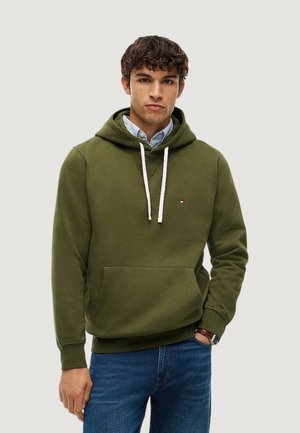 Ung mand iført en olivengrøn hættetrøje over en blåstribet skjorte og blå jeans, stående med den ene hånd i lommen mod en ensfarvet baggrund.