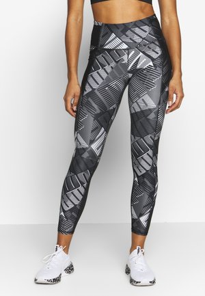Leggings à motifs noir et blanc avec des formes géométriques, taille haute et tissu extensible, associés à des chaussures de sport blanches.