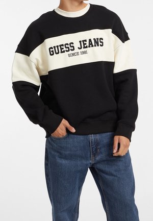 Person trägt schwarzen und weißen Pullover mit dem Text „GUESS JEANS SINCE 1981“ und blaue Jeans, Hand in der Tasche, vor weißem Hintergrund.