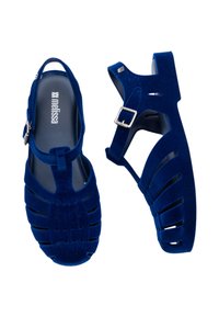 Niebieskie, aksamitne sandały z wycięciami, regulowanym paskiem wokół kostki i teksturowaną powierzchnią. Charakteryzują się płaską podeszwą i odkrytymi palcami.