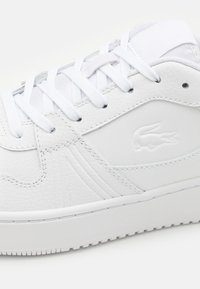 Hvite sneakers laget av teksturert lær, med en glatt overflate, flate snøringer og en subtil preget logo på siden.