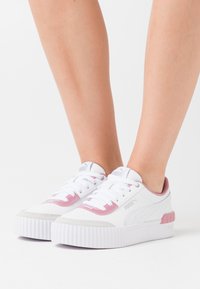 Baskets Puma blanches avec des accents roses portées sur des pieds devant un fond blanc uni, montrant des vues latérales et frontales.