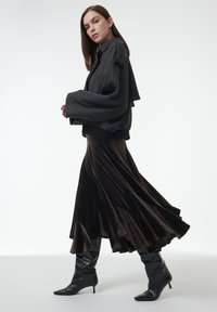 Chaqueta de gran tamaño en gris oscuro con mangas anchas, combinada con una falda de terciopelo marrón fluida y en capas. Botas de tacón alto negras con punta de aguja hasta la rodilla.