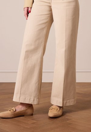 Beige weite Hosen aus strukturiertem Stoff, kombiniert mit hellen Slippern mit goldenen Hardware-Akzenten.