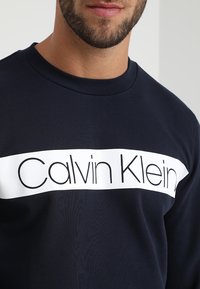 Námořnická modrá mikina vyrobená z bavlny, se bílým horizontálním pruhem s nápisem "Calvin Klein" vytištěným silnými černými písmeny přes hruď.