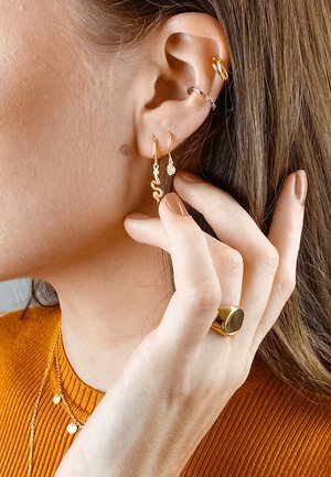 Nahaufnahme eines Frauenohrs mit mehreren goldenen Ohrringen, darunter eine Schlange, ein kleiner Anhänger und Ringe, die ihr Gesicht berühren. Sie trägt einen orangefarbenen Pullover.