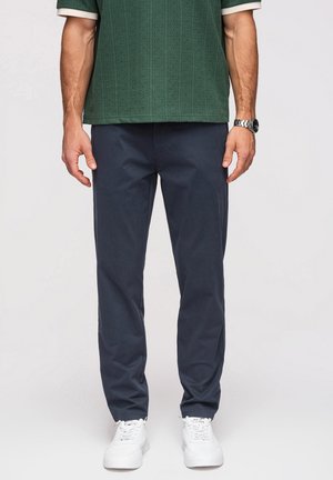 Homme portant un pantalon bleu marine foncé, des baskets blanches, une chemise verte texturée à manches courtes et une montre au poignet gauche, debout sur un fond uni.