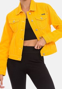 Yellow denim jacket with a classic collar, button closure, two chest pockets, and contrasting stitching, paired with a black top.