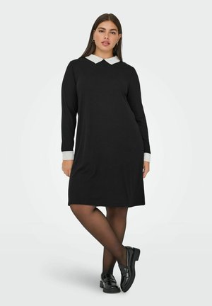 Must kleit pikkade varrukatega ja valge kraega, lõdva lõikega. Modell kannab musti sukkpükse ja musti mokkasiine.