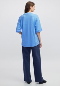 Světle modré oversized tričko s ohrnutými krátkými rukávy. V kombinaci s tmavě modrými kalhotami s širokými nohavicemi, vše na neutrálním pozadí.