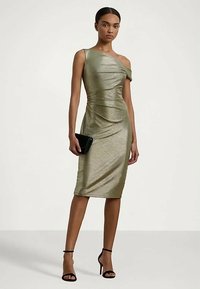 Γυναίκα που φοράει μεταλλικό ελαιόχρωμο φόρεμα ενός ώμου μέχρι το γόνατο, κρατώντας μια μαύρη τσάντα χειρός και φορώντας μαύρες ψηλοτάκουνες σάντες, στέκεται μπροστά σε λευκό φόντο.