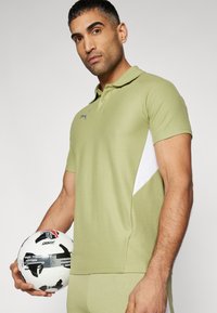 Oliven grøn polo shirt med en hvid sidestribe, klassisk krave, to-knap lukning og diskret logo detalje. Modellen holder en sort og hvid fodbold.