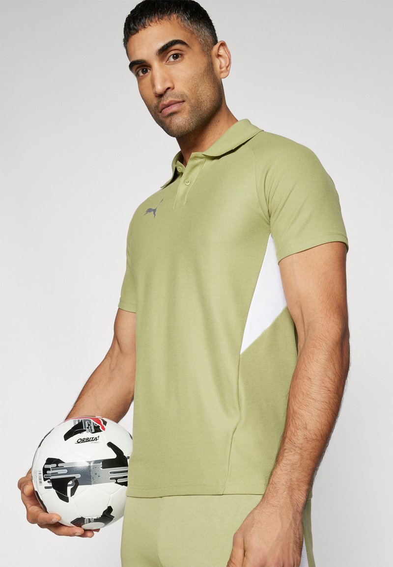 Olive green polo shirt with a white side panel, classic collar, two-button placket, and subtle logo detail. Model holds a black and white soccer ball.
