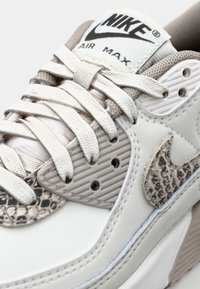 Nike Air Max kedas ar baltu ādas virsu, beidzīšu akcentiem, čūskas raksta detaļu sānos un teksturētām šņorēm.