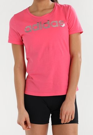 Femme portant un t-shirt Adidas rose avec logo argenté et un short noir, debout avec les bras détendus sur les côtés devant un fond uni clair.