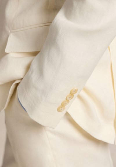 Šviesiai smėlio spalvos lininio kostiumo švarkelio rankovė su penkiais geltonais mygtukais, subtilios tekstūros ir mėlyna pamušalo detalė matoma rankogalyje.