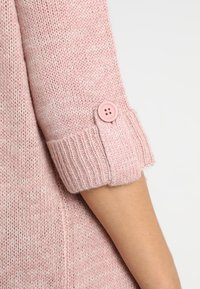 Różowa, dziana rękawica swetra podwinięta i zabezpieczona pasującym różowym guzikiem na jasnej skórze ręki, w tle na jednolitym tle.