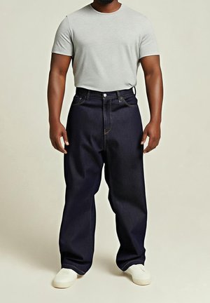 Man wearing plain light gray short-sleeve t-shirt, dark loose-fitting jeans, and white sneakers, standing against neutral background.