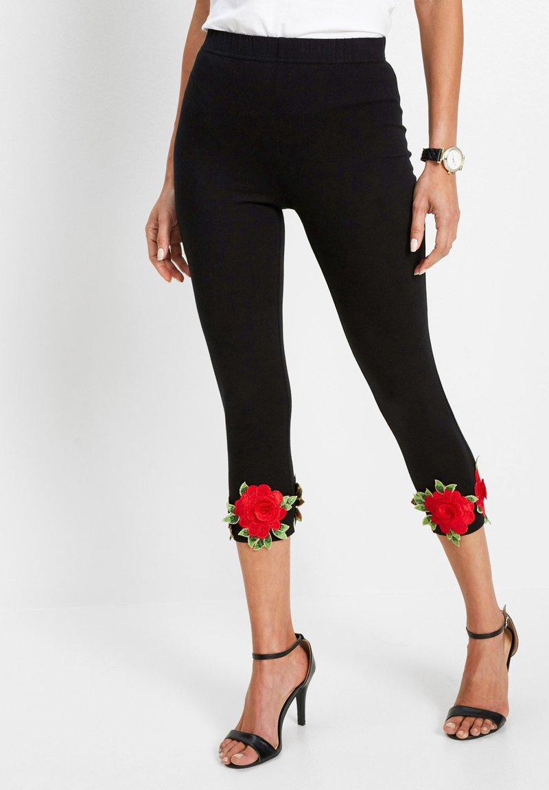 Donna che indossa leggings neri a vita alta con ricami di rose rosse vicino all'orlo, abbinati a sandali neri con tacco alto e una maglietta bianca.