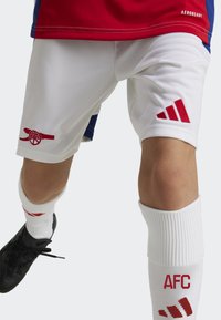 Vita sportshorts med röd Arsenal-logga, parat med vita strumpor med röda detaljer och Adidas-logga. Tyget verkar slätt och syntetiskt.