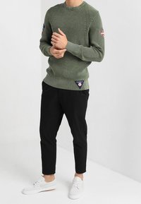 Man in een olijfgroen gebreid sweater met patches, zwarte slim-fit broek en witte veterschoenen staat tegen een plain witte achtergrond.