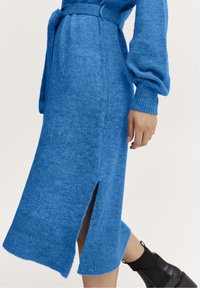 Robe tricotée bleue avec des manches longues, des poignets côtelés et une fente sur le côté. Elle dispose d'une ceinture assortie et d'un tissu texturé. Longueur chevilles.