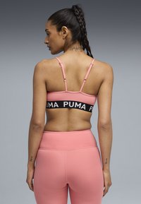Γυναίκα με πλεγμένα μαλλιά που φοράει ροζ αθλητικό σουτιέν με μαύρη ταινία PUMA και ροζ κολάν που ταιριάζουν, εμφανιζόμενη από πίσω μπροστά σε γκρι φόντο.