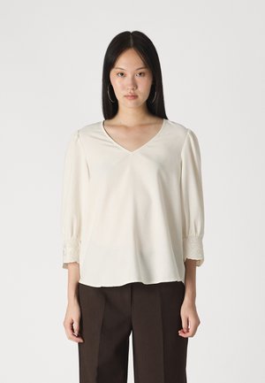 Cream blouse with a V-neck, puffed sleeves, and lace detailing at the cuffs. Smooth texture, loose fit, paired with dark trousers.