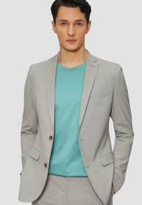 Blazer grigio chiaro con trama sottile e due bottoni dorati, abbinato a una t-shirt a girocollo turchese, ambientato su uno sfondo semplice.