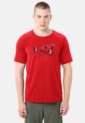 Uomo con t-shirt rossa a maniche corte con una grafica da escursionismo e pantaloni verdi, in piedi contro uno sfondo chiaro e uniforme.