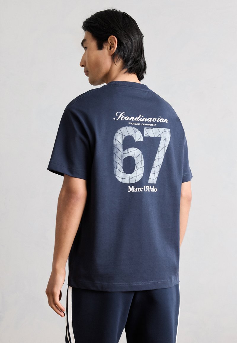 Άνδρας με μπλε ναυτικό t-shirt με το κείμενο «Scandinavian 67 Marc O'Polo» και σχέδιο με δίχτυ, στέκεται πλαγίως σε μονόχρωμο φόντο.