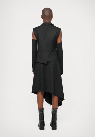 Melns vestais ar plecu izgriezumiem, kombinēts ar asimetrisku svārku. Apģērbs ir šūts pēc pasūtījuma un tam ir matēta tekstūra. Izskatu papildina melni zābaki.