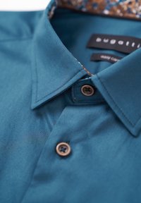 Türkisfarbiges Hemd mit Knöpfen, das eine glatte Oberfläche, ein kontrastierendes, gemustertes Kragenfutter und braune Knöpfe aufweist. Markenetikett sichtbar im Kragen.