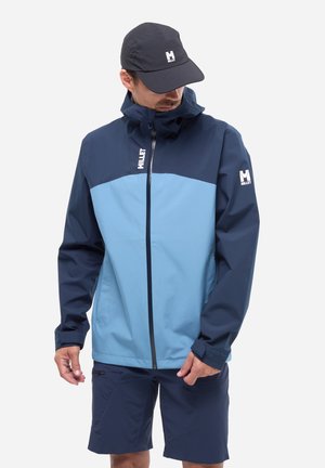 Man wearing a two-tone blue Millet jacket with a front zipper, navy shorts, and a black cap, looking down and adjusting his jacket.