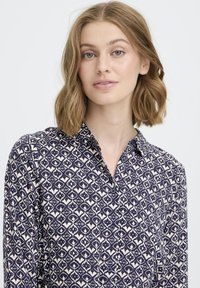 Moteris su pečių ilgio banguotais šviesiais plaukais, dėvinti tamsiai mėlyną ir baltą raštuotą marškinėlį su sagomis, prieš šviesų vientisą foną.