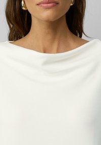 Witte top met een gedrapeerde halslijn, gemaakt van gladde stof, met een subtiele textuur. Aangehechte gouden hoepeloren zichtbaar op de voorgrond.