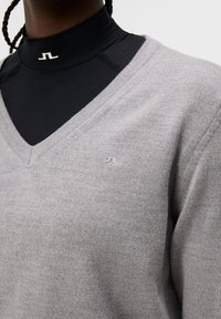 Grå v-ringad tröja i mjukt stickat material, med en subtil logodetalj på bröstet och lager p å lager över en svart högkragad topp.