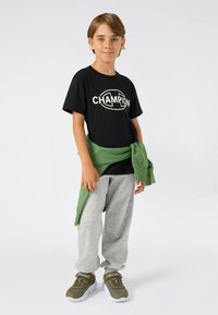 Berniukas stovi vilkėdamas juodą „Champion“ marškinėlį, pilkas sportines kelnes, žalias sportines batų ir žalią džemperį surištą aplink liemenį prieš vienodą foną.