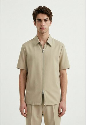 Unge mann iført beige kortermet skjorte med glidelås foran og matchende bukser, stående mot en ensfarget lys bakgrunn.